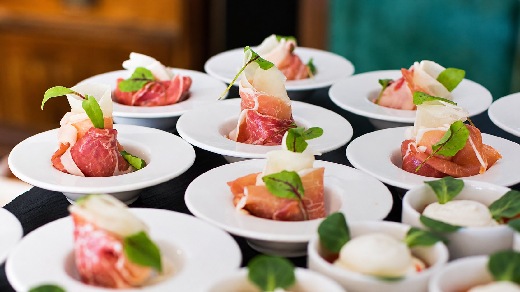 Plusieurs petites assiettes blanches creuses présentent du jambon cru roulé et de la poire, décorés de feuilles vertes, avec des bols de mozzarella au premier plan.