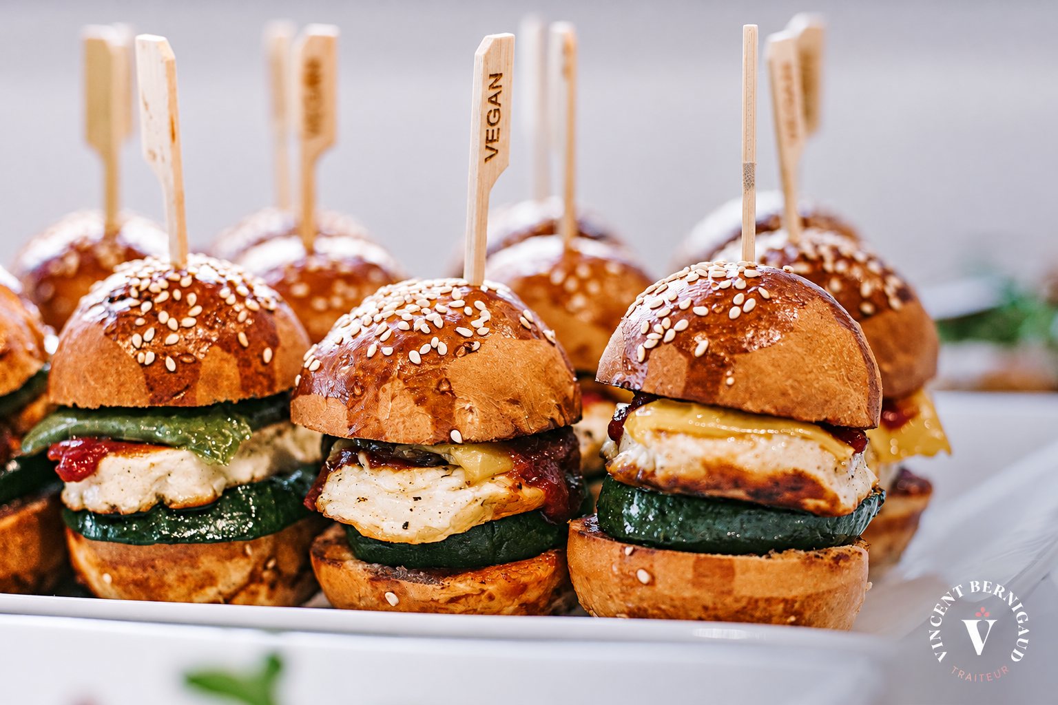 Plateau de mini burgers briochés au sésame, garnis de poulet, courgette et fromage, avec une pique indiquant VEGAN.