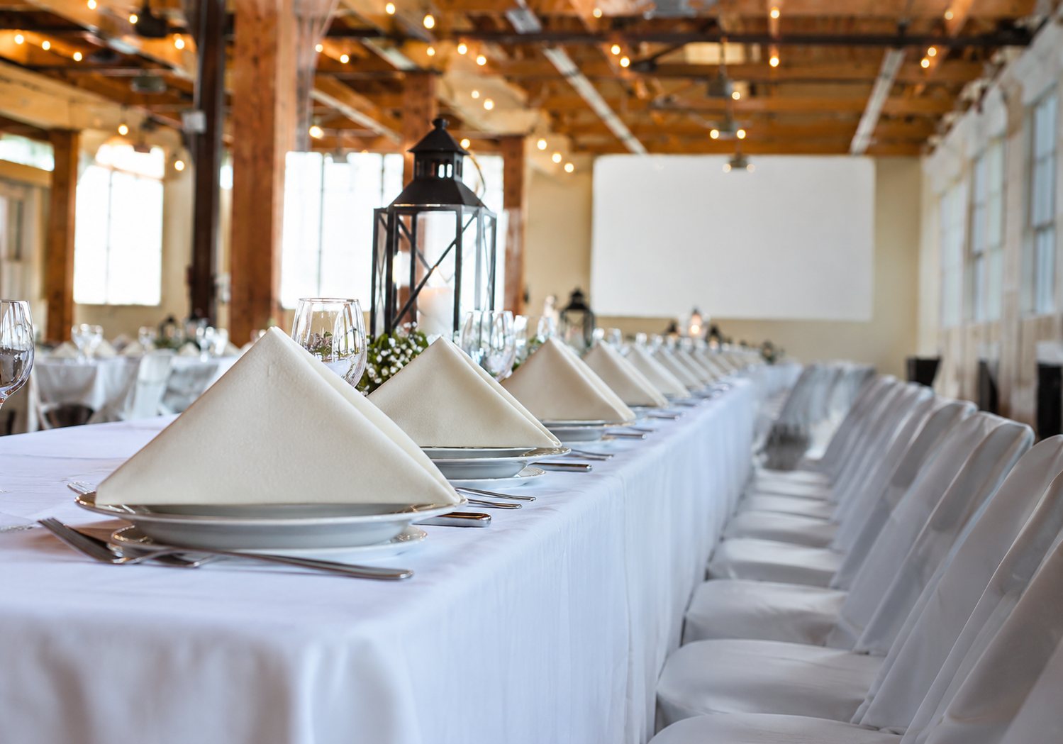 Salle de réception avec de longues tables couvertes de nappes blanches et de serviettes pliées, prêtes pour accueillir des convives.